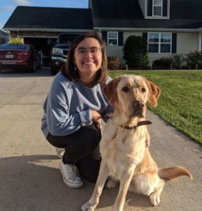 Brooke crouches, smiling, on a driveway in front of a two-story house, She is holding Sampson's leash as he sits next to her