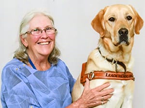 Yellow lab/golden retriever in a Leader Dog harness sits next to a smiling woman with graying hair and glasses.