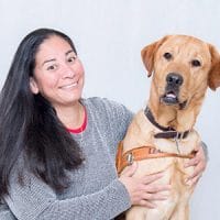 Jennifer smiles with both her arms around yellow lab Mosby. Mosby is seated next to her in Leader Dog harness