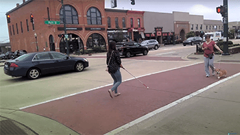 A woman with a white cane crosses a street on a brick crosswalk. There is traffic in the street behind her.