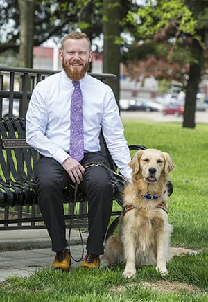 David Locklin sits on a bench outdoors wearing a white collared shirt and dark pants with a purple tie. Next to him on the ground is a sitting golden retriever and David has his hand on its back
