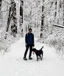 Justin, man in dark colored, warm clothing, stands on a snow-covered trail in the woods with a black lab at his side. He's smiling at the camera