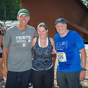 A woman and two men, all smiling, stand with their arms around each other. They are wearing athletic gear and one man has a Bark & Brew 5K race bib on the front of his shirt