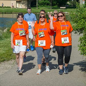 A group of six women and men wearing orange Bark & Brew 5K race shirts and bibs with athletic gear walk on a cement pathway, smiling