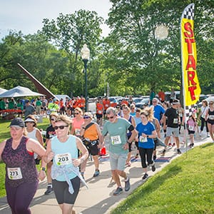 A large group of people wearing Bark & Brew 5K bibs and athletic gear run down a sidewalk outdoors near a large vertical banner that says START