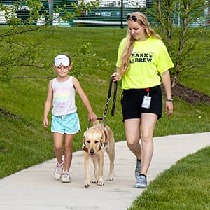 Bark & Brew blindfold walks Heidi A blonde woman walks along a sidewalk holding the leash of a yellow lab. A young girl is walking next to the dog holding its harness. She has a blindfold pulled up over her eyes.