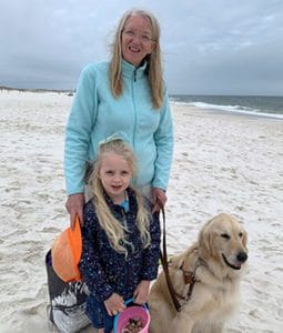 A woman, young girl and golden retriever are on a sandy beach with water and waves in the background. The woman is standing in the back. She has long blond hair, round, wire-rimmed glasses and a smile on her face. She is wearing a turquoise fleece jacket and is holding the dog’s brown leather leash in her left hand. The young girl in front has long blond hair partially pulled back with a ribbon. She is wearing a navy-blue skirt and jacket and is holding a pink and blue plastic bucket containing shells. The dog is wearing a leather LDB harness and calmly sits next to the girl.