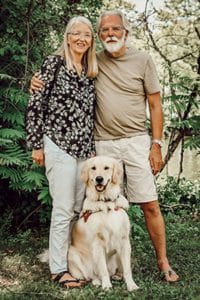 A woman and man are standing with a golden retriever wearing a harness sitting in front of them on grass. The background is a variety of trees and a river. The woman is smiling, she has long blond hair and round, wire-rimmed glasses. She is wearing a long-sleeve black blouse with a tan floral/leaf patten and khaki pants. The man has grey hair and a full white beard and moustache. He is wearing round glasses, a tan short-sleeve t-shirt and khaki shorts. The dog has very light-yellow fur, dark eyes and a dark nose. His mouth is slightly open, and you can see a black spot on his light pink tongue.