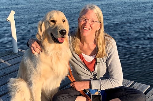 A woman and a golden retriever are sitting on a wooden dock with a rippling blue lake in the background. The woman is smiling and sitting cross-legged with her right arm around the dog’s shoulders, she holds the dog’s brown leather leash in her left hand. She has long, straight blond hair and round wire glasses; she is wearing denim capri’s and a white, red and light grey top. The dog’s tongue is hanging out and has a black spot in the middle. He has long light tan fur and a very black nose.