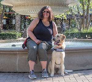 A smiling woman in black tank top and gray capris sits on the edge of a fountain next to a golden retriever in blue Leader Dog vest.