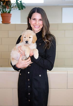 Alex is in a black button-up dress, smiling and holding a young golden retriever puppy