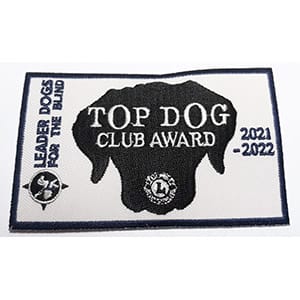 A white rectangular patch with a dark blue border. In the center of the patch is a dark blue silhouette of a dog head and the Lions International logo. The patch features embroidered text, "Leader Dogs for the Blind, Top Dog Club Award 2020-2021