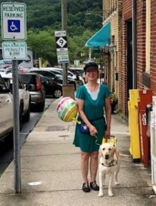 A smiling woman wearing a black hat and green dress is standing on a sidewalk with the road on her right and red brick buildings on her left. She is holding the handle of a yellow Labrador/golden retriever cross in her left hand and a balloon in her right hand. Between the woman and the road is a street sign with “Reserved Parking” and a blue square with a white graphic of a person in a wheelchair.