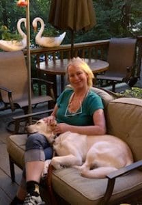 A woman is sitting on a loveseat on a patio. She is wearing a green t-shirt, jean capri’s and had white glasses hanging from a cord around her neck. A yellow dog is lying on the loveseat next to the woman with its head on her lap. The dog appears to be asleep.
