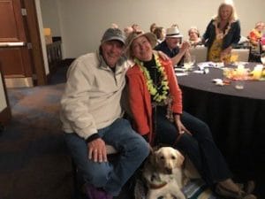 A man and a woman, both smiling toward the camera, are seated at a large round table in a hall. There are people in the background seated at their table and other tables. The man is wearing a baseball cap and cream-colored fleece jacket. The woman is wearing a floppy hat, black shirt and pants, orange jacket and a lei around her neck. At their feet is a yellow dog lying on the floor.