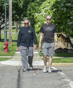 A man in sunglasses walking in a crosswalk with a white cane on a sunny day