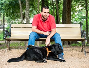 A dark-haired man sits on an outdoor bench with a black lab in Leader Dog harness lying across his feet