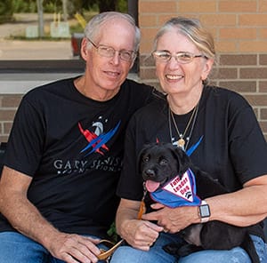 The Gimbles, both wearing black t-shirts with the Gary Sinise Foundation logo, sit next to each other, smiling. Linda Gimble has a black lab puppy, Jenny, on her lap.