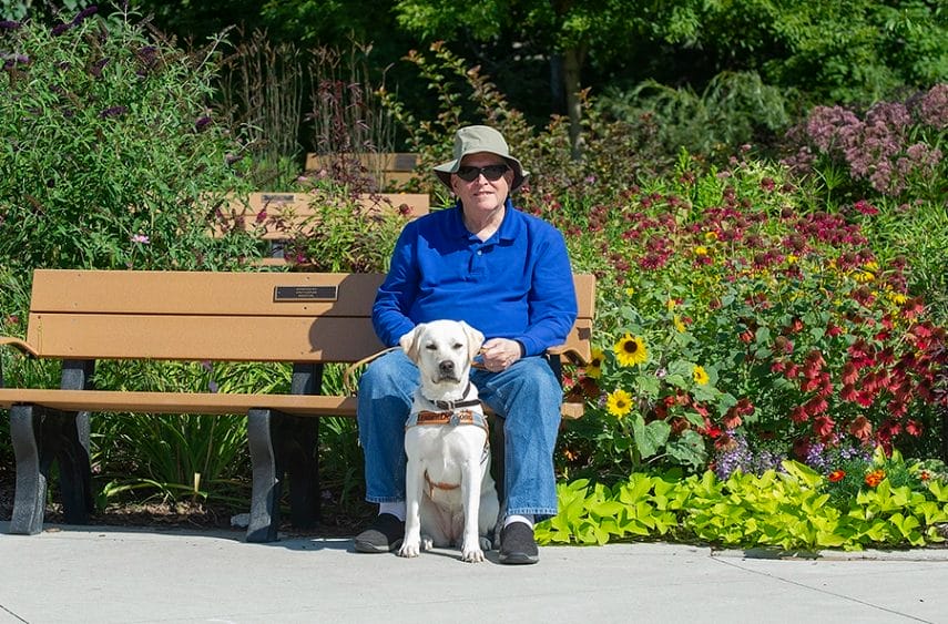 Mark sits on a park bench with flowers and greenery behind him. Yellow lab Leader Dog Izzy sits in front of him on the cement in her harness.