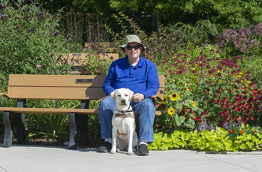 Mark sits on a park bench with flowers and greenery behind him. Yellow lab Leader Dog Izzy sits in front of him on the cement in her harness.