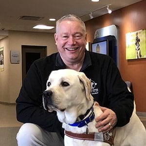 Jim is in a black long sleeve shirt and light colored pants kneeling and smiling next to a yellow lab in guide dog harness