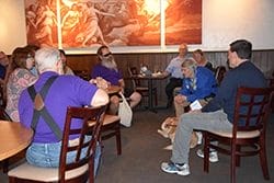The interior of a restaurant with men and women turned to face one woman, Pauline, who is speaking. Pauline is leaning forward with her elbows resting on her knees. A yellow Labrador retriever, Satch, is wearing a brown leather harness and lying at Pauline’s feet.