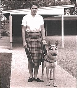 A black and white photo of a young woman wearing a white short-sleeve cotton blouse and a pleated plaid skirt and flat shoes. In her left hand is a harness handle attached to a light tan German shepherd that is standing by the woman’s side. Behind them is a large awning over an area covered in pea gravel.
