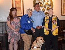Inside a restaurant three women, a man and a yellow Labrador retriever stand side by side. From left to right are District 42 Governor Lion Sally Schroth wearing a grey and peach dress; Lion Pauline Ulrey wearing khaki pants and a blue shirt, in her left hand is a leash connected to the dog that is wearing a brown leather harness; Incoming Narragansett Lions Club President Lion Eric Menke wearing a blue button down shirt with the Lions emblem over the chest pocket; and Lion Alescia “AJ” Johns wearing dark pants, a dark shirt and a bright gold Lions vest with many patches and pins.