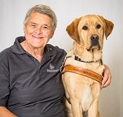 A woman with short, grey hair wearing a grey polo shirt with the old Leader Dog logo and khaki pants. She has a large smile on her face and her left arm is hugging a yellow Labrador retriever that is wearing a brown leather Leader Dog harness. The dog is light, golden tan with darker gold ears.