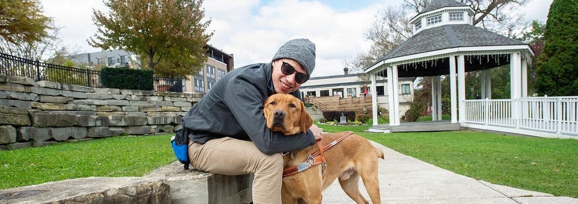 A man in a gray beanine and jacket with sunglasses smiles while hugging a gold-colored yellow lab with its eyes closed next to him on a sidewwalk. The man is sitting on a low wall