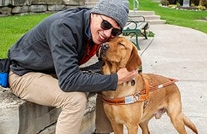 A man in a gray beanine and jacket with sunglasses smiles while hugging a gold-colored yellow lab with its eyes closed next to him on a sidewwalk. The man is sitting on a low wall