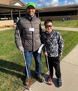 A smiling man and boy stand outdoors in puffy coats. Each is holding a white cane.