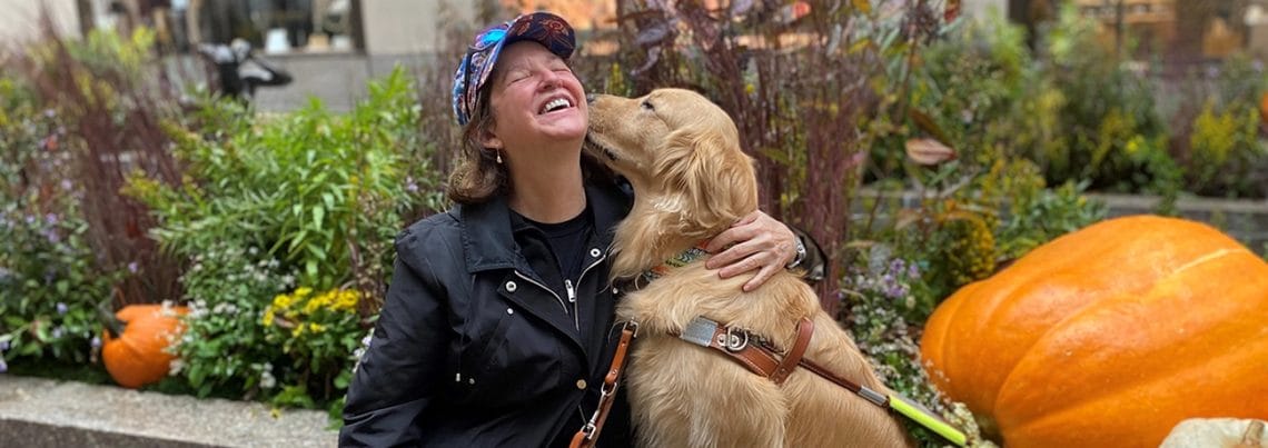 Smiling woman with closed eyes leans her head back while getting a kiss from a golden retriever in Leader Dog harness sitting next to her. They are outdoors near some green and brown plants and decorative pumpkins.