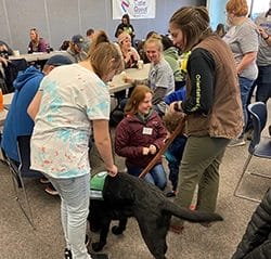 Room full of people sitting and talking with a girl petting a black lab in a green vest in the foreground. In the background is a sign on the wall saying "Cane Quest"