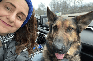 A closeup selfie of a woman, Jill, and a German shepherd, Leader Dog Hannah, in the front passenger seat of a car. Jill is smiling and wearing a winter coat, scarf and knit headband. Hannah has her pink tongue slightly hanging out of her mouth. In the background is a children play structure and a line of trees without leaves.