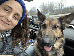 A closeup selfie of a woman, Jill, and a German shepherd, Leader Dog Hannah, in the front passenger seat of a car. Jill is smiling and wearing a winter coat, scarf and knit headband. Hannah has her pink tongue slightly hanging out of her mouth. In the background is a children play structure and a line of trees without leaves.