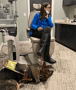 A woman, Jill, is sitting in a large chair in an optometrist office. Next to her is a large piece of medical equipment. On the floor at her feet is a large, dark German shepherd, Leader Dog Hannah, lying down. Hannah is wearing a leather guide dog harness with a sign with the words “Do not pet me, I’m working.”