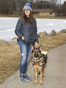 A woman, Jill, standing on a paved walkway in a park with a German shepherd, Leader Dog Hannah, in harness standing next to her. They are facing the camera. Jill is smiling and wearing a Grand Valley State University knit hat, a black winter coat and a dog treat bag on her hip. Hannah has large, pointed ears, her face is mostly black with some tan, and her front legs are all tan. There is a frozen pond in the background.