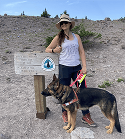 A woman, Jill, is standing in a rustic area that is mainly dirt with some shrubs and small trees. She is smiling and resting her right arm on a sign with the words “Pacific Crest National Scenic Trail,” and she is giving a “thumbs up.” Jill is wearing a hat with a large, floppy brim, sunglasses, hiking shoes, black jogging pants and a grey tank top with the words “You, My Dear, Are Brave.” She is holding onto the guide dog harness handle of a German Shepherd, Leader Dog Hannah.