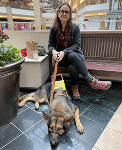 A woman, Jill, is sitting on a bench in the middle of a large shopping mall. A large German shepherd, Leader Dog Hannah, is lying on the tile floor at Jill’s feet. Jill is wearing dark tortoise shell glasses and is casually dressed. Hannah is wearing a brown leather harness and a leather leash which Jill is holding.