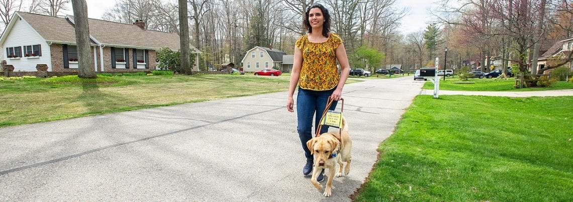 A smiling woman with curly dark hair walks on the edge of a paved road with a yellow lab Leader Dog in harness. The street is lined by suburban houses and bare trees.
