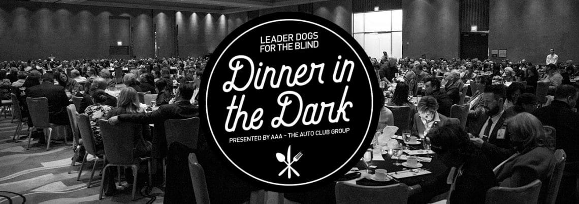 Black and white photo showing a large room of people at round tables in a banquet room. Overlaid is a black and white Dinner in the Dark logo