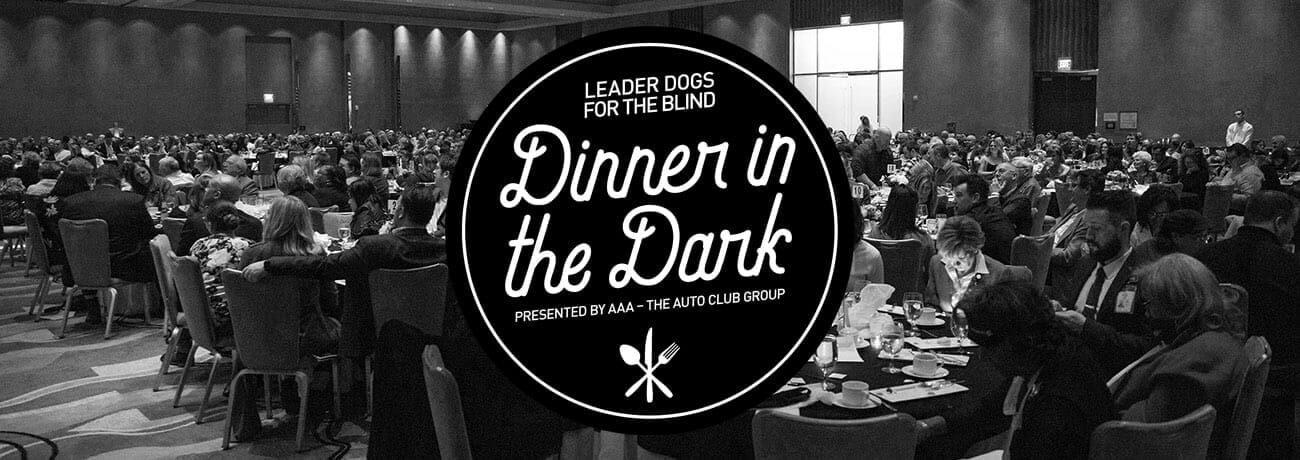 Black and white photo showing a large room of people at round tables in a banquet room. Overlaid is a black and white Dinner in the Dark logo