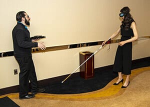 A blindfolded woman walks with a white cane in a carpeted room while a man gestures while speaking to her