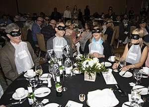 A group of people with black blindfolds on sit around a table with a black tablecloth and place settings. Other tables of people in blindfolds are behind them
