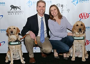 A man in a suit coat and tie and a woman in a lilac gray blouse smile while crouching next to two yellow labs wearing Canine Ambassador vests