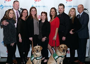 A group several men and six women in dressy clothes stand smiling with two yellow labs in green Canine Ambassador vests