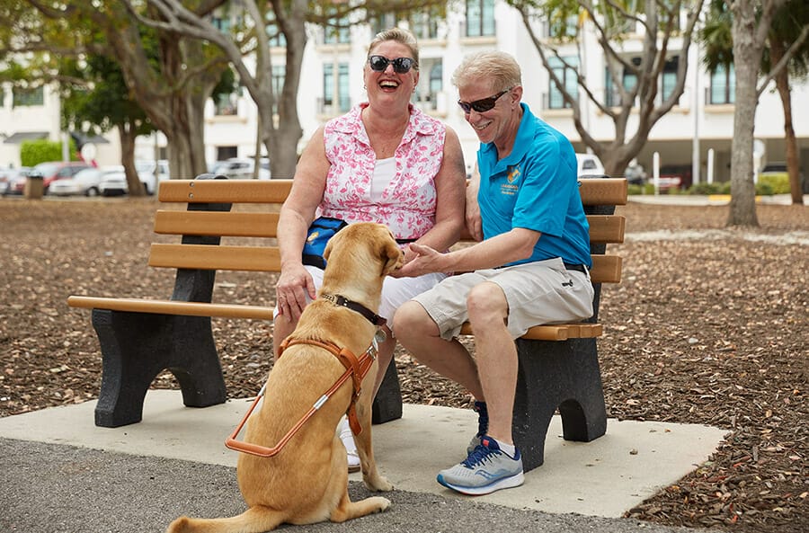 Keith, wearing sunglasses, khaki shorts and a blue Leader Dog polo, sits smiling next to a woman in sunglasses and a flowery top. The woman has her mouth in a wide open smile as if laughing. A yellow lab in harness sits near their feet looking at them.