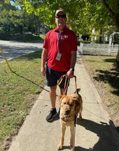 Joey, in dark shorts, red polo and khaki cap, stands on a grass-lined sidewalk with a yellow lab in Leader Dog harness slightly in front of him on his left.