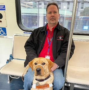 Joey sits on a train seat with Tucker sitting at his feet in Leader Dog harness.